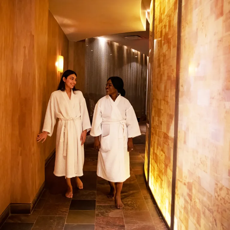 Two women in white bathrobes walking down Elements Spa hallway.
