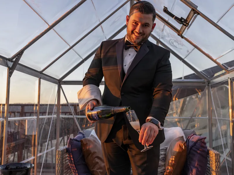 Waiter, dressed in a black suit,  pouring champagne while standing in the champagne chalet. 